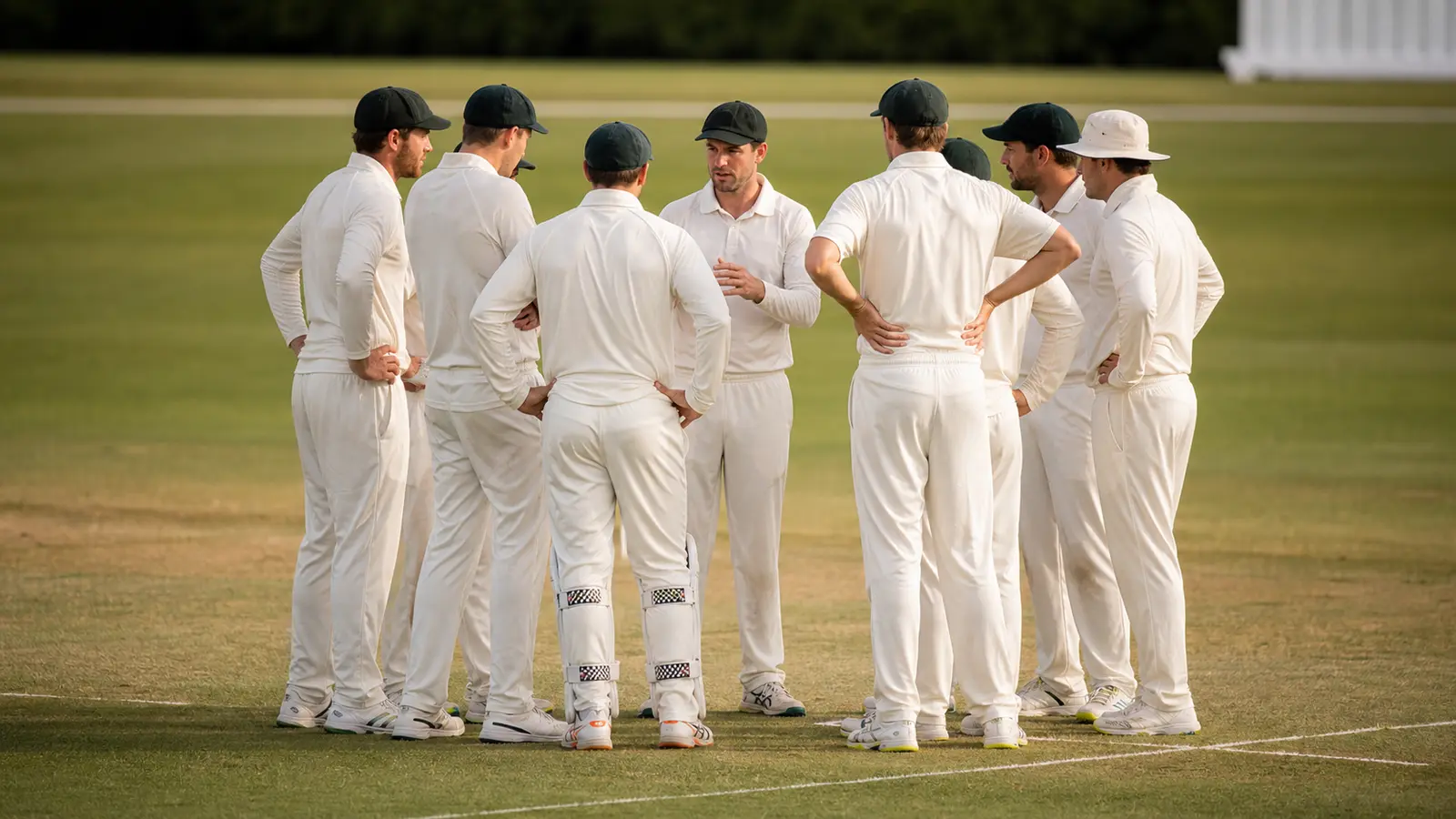 Cricket team in white kits gathered with the captain on the grass pitch during a tactical break in play